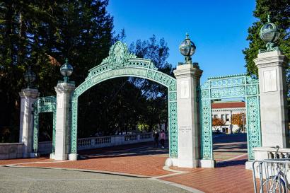 A view of the green and white Sather Gate, the red brick pathway, and a few trees all backdropped by a bright blue sky.
