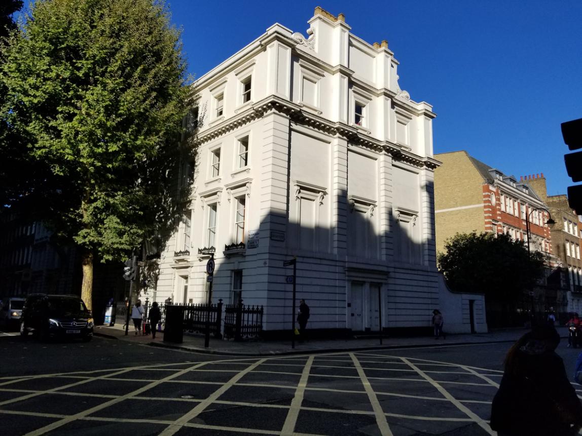 A multi-story white 18th-century Georgian building stands at the intersection of two streets. A large tree stands to the left of the building.