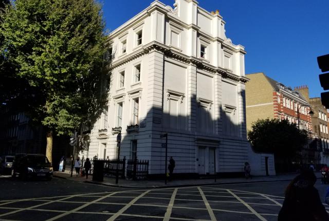 A multi-story white 18th-century Georgian building stands at the intersection of two streets. A large tree stands to the left of the building.