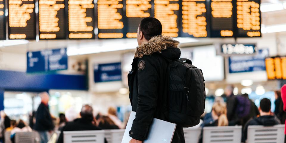 Man standing with backpack and laptop in hand looking at departure board at the airport.